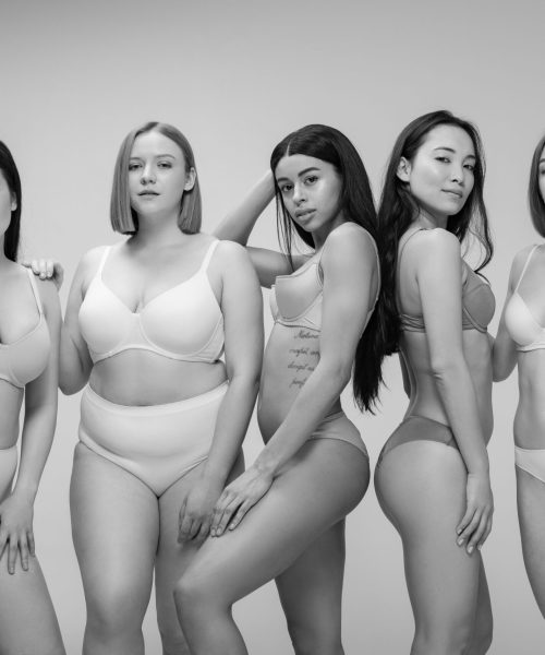 five smiling multiethnic girls smiling and posing at camera isolated on grey, body positivity concept